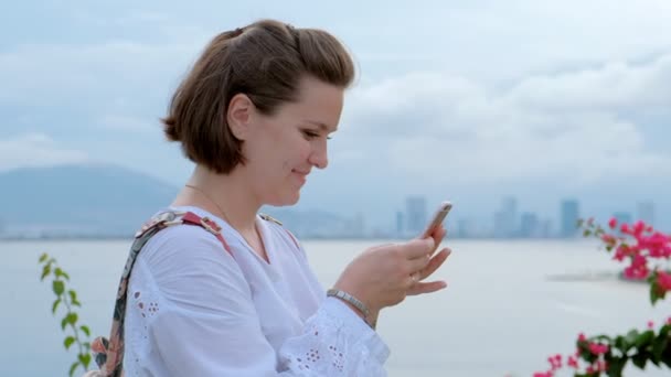 Un primer plano de una hermosa joven escribiendo en su teléfono inteligente con un océano y paisaje urbano en el fondo — Vídeo de stock