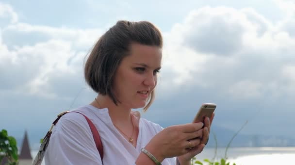 Uma foto lateral de uma bela jovem sorridente digitando em seu smartphone com um oceano e paisagem urbana no fundo — Vídeo de Stock