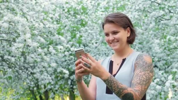 Linda mujer de cabello castaño joven está charlando con el teléfono inteligente en el jardín de manzanos en flor en el día soleado. estilo de vida emociones sonrientes. refrigeración al aire libre — Vídeo de stock