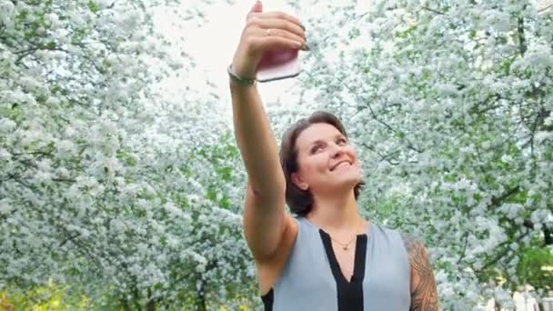 Joven linda mujer tatuada está tomando selfie en el teléfono celular en el huerto de manzanas en flor — Vídeos de Stock