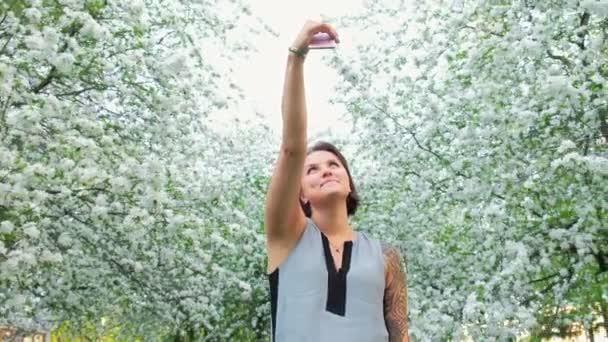 Young cute tattooed woman is taking selfie on cellphone in blooming apple orchard