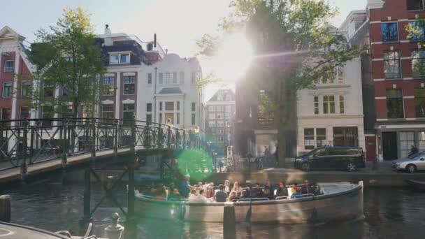 Amesterdam, Nederland, mei 2018: Schilderachtige landschap - een boot met toeristen zwemt onder de brug langs een van de grachten van de stad. Via de oude huizen de zon schijnt. — Stockvideo