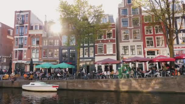 Amesterdam, Pays-Bas, mai 2018 : La rue pittoresque d'Amsterdam où les drogues légères sont légalisées. Les gens se reposent dans les cafés de la rue, le soleil couchant brille à travers les maisons — Video