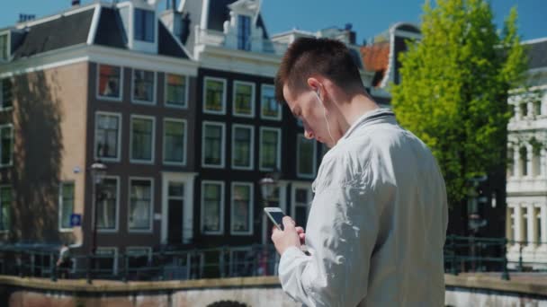 Un adolescente escucha música en los auriculares, tiene un teléfono inteligente en la mano. Está cerca del puente que cruza el canal en Ámsterdam. Turismo y estudio en los Países Bajos — Vídeo de stock