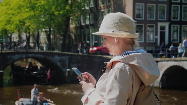 Een toeristische vrouw maakt gebruik van een smartphone in het centrum van Amsterdam. Op de achtergrond, het intensieve verkeer van boten, voetgangers en fietsers. — Stockvideo