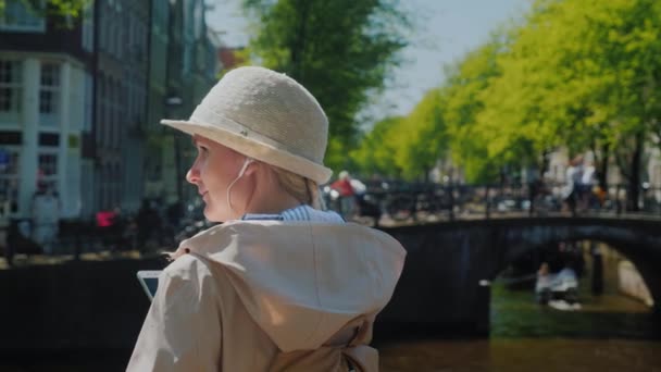 Dynamisches Leben der Stadt. eine frau mit kopfhörer und smartphone in der hand, im hintergrund eine intensive bewegung von radfahrern, fußgängern und schiffen auf dem kanal von amsterdam — Stockvideo