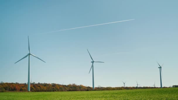 Flera vindkraftverk. I förgrunden finns ett grönt gräs, i bakgrunden ett plan flyger på himlen — Stockvideo