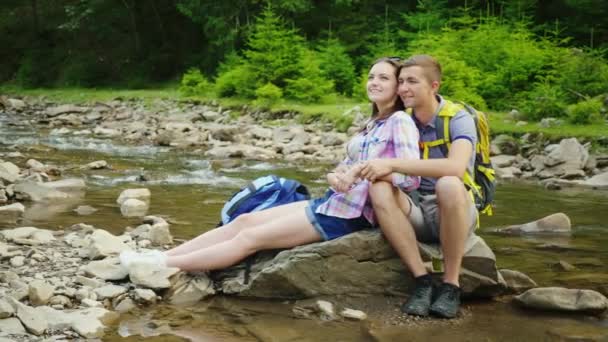 Turista hátizsákok-val egy szerető pár nyugszik, egy festői helyen, egy hegyi patak mellett. Ölelés, és egy rock ül. Kellemes Ünnepeket