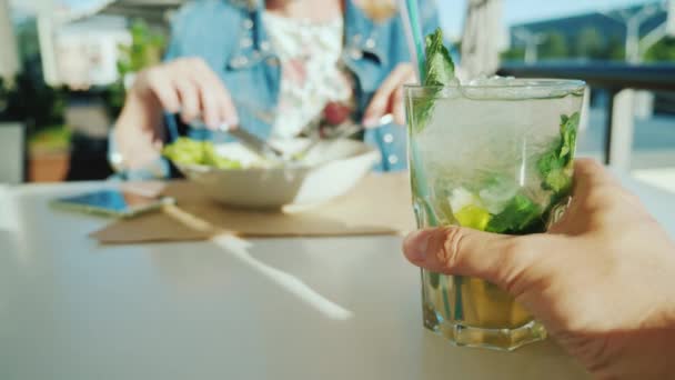 Ein Paar ruht sich in einem Café aus. Ich-Perspektive - ein Mann hält ein Glas mit einem Cocktail in der Hand, eine Frau im Hintergrund isst einen Salat — Stockvideo