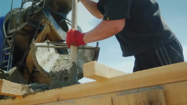 Рабочий берет бетон из промышленного смесителя, работает как лопата. Тяжелый ручной труд на стройке. Снимок с низким углом — стоковое видео