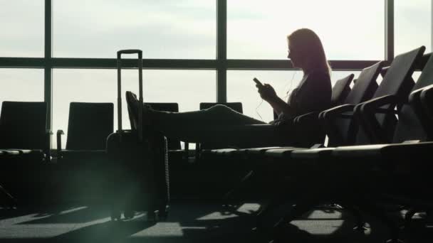 Una mujer está sentada en la terminal del aeropuerto internacional, esperando su vuelo. Utiliza un teléfono inteligente — Vídeos de Stock