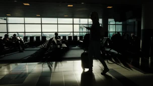 ホイールと彼女の手で携帯電話にバッグを持つ女性は、空港ターミナルを歩いています。大きな窓の背景 Silhouet — ストック動画
