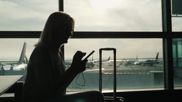 女性は、大きなターミナル ウィンドウで彼女の飛行を待っています。携帯電話を楽しんでいます — ストック動画