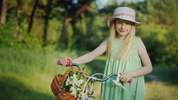 Retrato de una chica rubia con una bicicleta. Con su cesta de flores silvestres. Mira la cámara. — Vídeo de stock