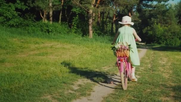 Una ragazza con un vestito verde estivo sta cavalcando lungo un sentiero nella foresta. Porta un cesto di fiori. Concetto vacanza estiva attiva — Video Stock