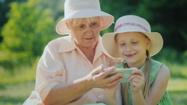 Бабушка и внучка используют смартфон вместе. Отдохните в парке в летний день — стоковое видео