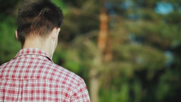 Un adolescente cammina nel parco ascoltando musica con le cuffie. Vista posteriore, video con bassa profondità di campo — Video Stock