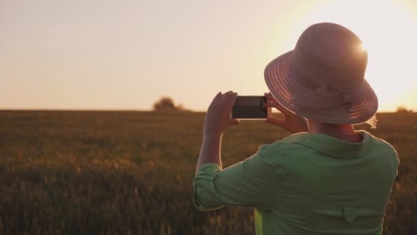 Женщина в шляпе фотографирует красивый закат над пшеничным полем — стоковое видео