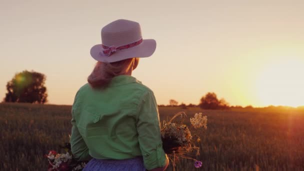 Een romantische vrouw met een hoed en een boeket van wilde bloemen bewondert de zonsondergang over het tarweveld. Achteraanzicht — Stockvideo