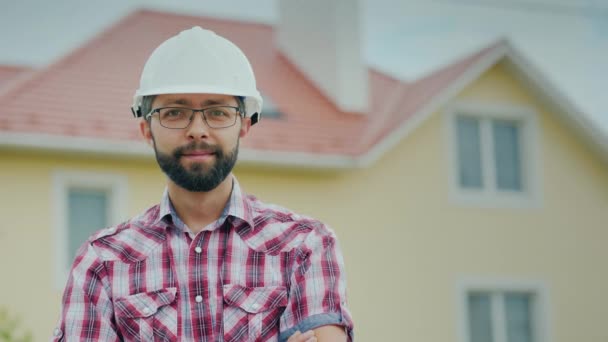 Retrato de una brigada en un casco en el fondo de una casa de campo moderna. Profesional en la construcción de un concepto — Vídeos de Stock