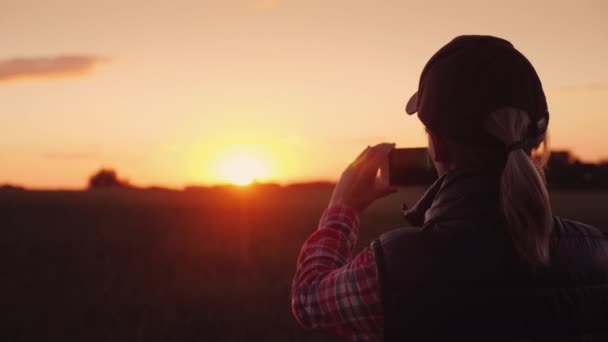 Mujer joven agricultora fotografiando una hermosa puesta de sol sobre un campo de trigo — Vídeo de stock