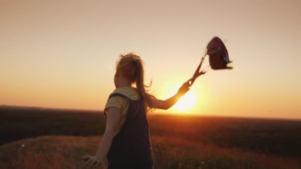 Uma criança feliz está acenando uma mochila e correndo em direção ao sol. Conceito de férias de verão — Vídeo de Stock