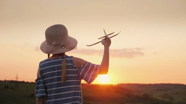 Ein Mädchen mit Zöpfen und Hut spielt bei Sonnenuntergang mit einem hölzernen Flugzeug. Der Traum vom Fernverkehr ist ein Konzept — Stockfoto