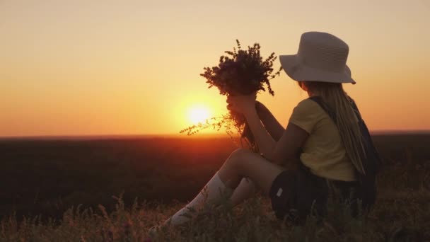 Una chica con un ramo de flores silvestres en sus manos está mirando una hermosa puesta de sol. Se sienta en una colina en un lugar pintoresco — Vídeos de Stock