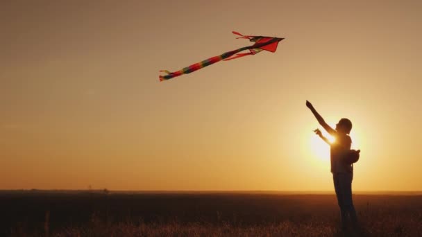 Nő egy gyermek hátizsák, háta mögött fut a kite a naplemente. Álom és visszatérés a gyermekkor fogalma — Stock videók