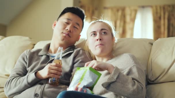 Genç çift birlikte televizyon izliyor. Bir adam bira içiyor, bir kadın bir paket aperatifler tutuyor — Stok video