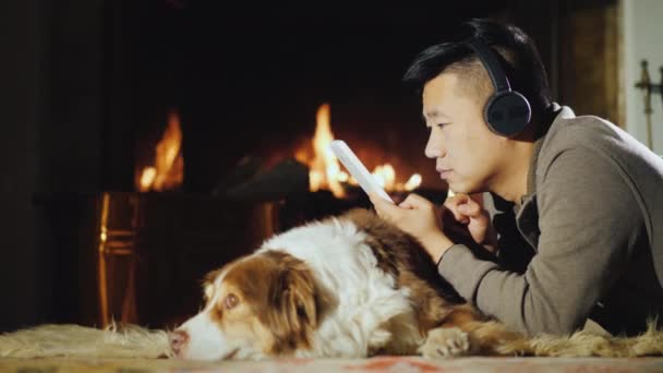 Un joven asiático usa una tableta cerca de la chimenea. Muy cerca está su perro. Calidez y confort en el concepto de la casa — Vídeos de Stock