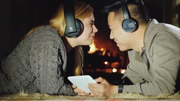 Una pareja joven se está divirtiendo en casa junto a la chimenea. Disfrute de una tableta, escuchar música en los auriculares — Vídeo de stock