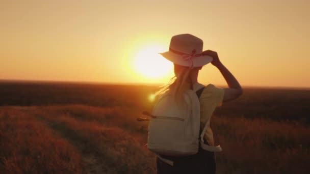 Rapariga engraçada de chapéu de palha desce a colina ao pôr-do-sol. Segura o chapéu com as mãos. Leto e o conceito de férias — Vídeo de Stock