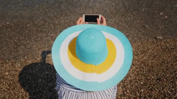 Una mujer en un vestido de verano y sombrero está fotografiando el surf. Mujer en concepto de vacaciones — Vídeos de Stock