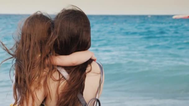 Молодая мать с каштановыми волосами обнимает свою маленькую дочь с длинными струящимися волосами у синего моря — стоковое видео