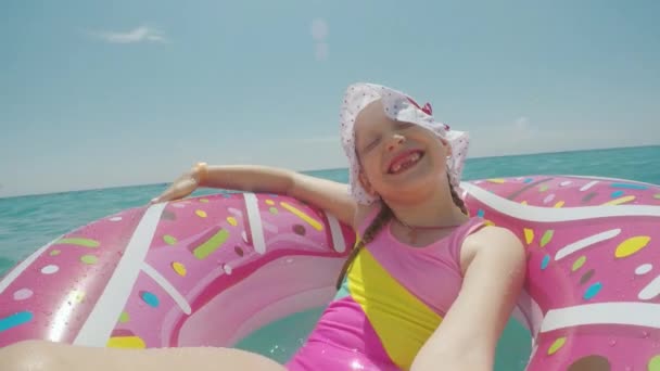 Una niña en un sombrero fresco y un bikini brillante está montando en un círculo y haciendo una selfie, sonriendo a la cámara — Vídeo de stock