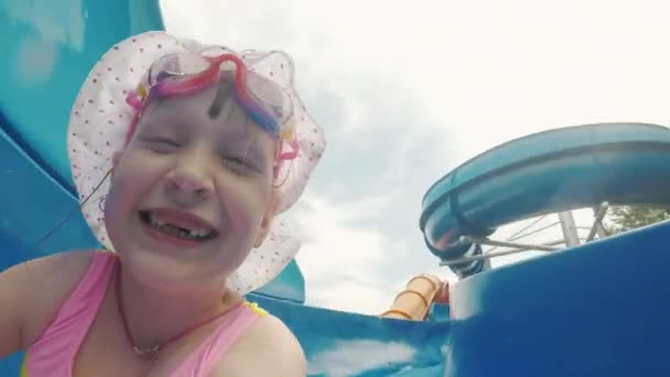 Ein cooles Mädchen in rosa Badeanzug und pinkfarbener Brille steigt von der Wasserrutsche im Wasserpark herunter, macht fröhlich ein Selfie in die Kamera — Stockvideo