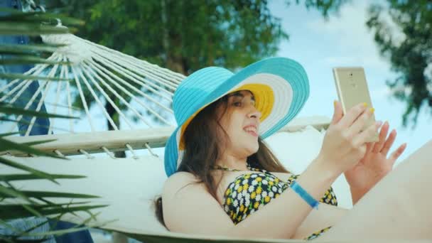 A cute brunette in a wide-brimmed hat and in a bikini enjoys relaxing in the seaside resort, reading sms on the phone — Stock Video