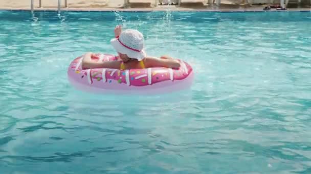 A funny little girl in a panama skates on an inflatable circle in a large pool in a hotel on the sea. Rear view — Stock Video