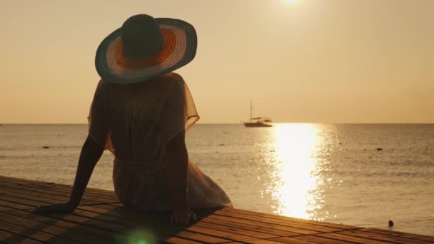 Mladá žena se setká s východem slunce na molu. Sedí a dívá se na slunce a loď v moři. Sny a romantický koncept — Stock video