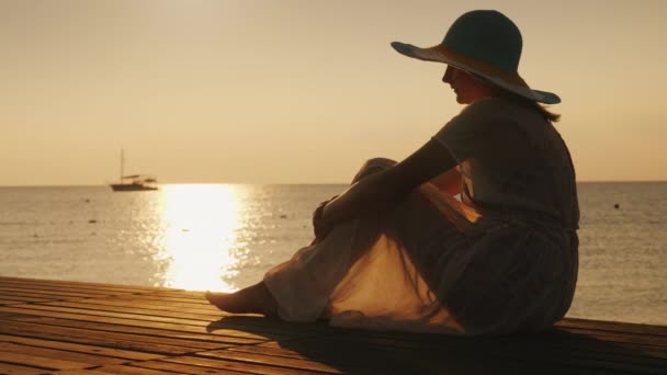 Zijaanzicht: Een vrouw zit op een houten pier, ontmoet de dageraad aan zee. In de verte is een schip te zien. Dromen en reizen concept — Stockvideo