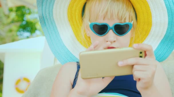 Uma menina loira com um chapéu e óculos de sol usa um smartphone. Ele está descansando no leito do sol. Férias com conceito de crianças — Vídeo de Stock