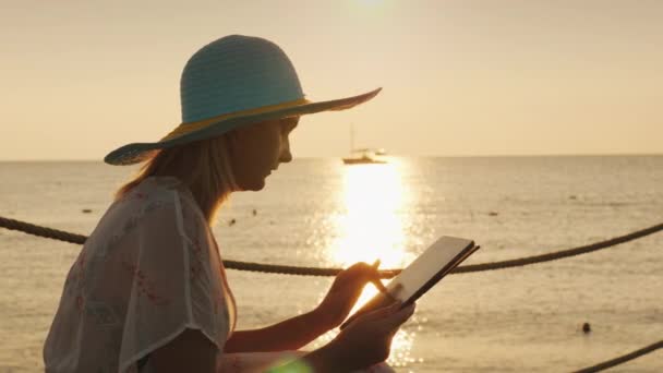 A woman uses a tablet on vacation. Sits against the background of the rising sun above the sea. Always in touch concept — Stock Video