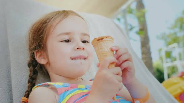 A happy little girl enjoys summer vacations, lies on a couch at sea and eats a delicious ice cream — Stock Video