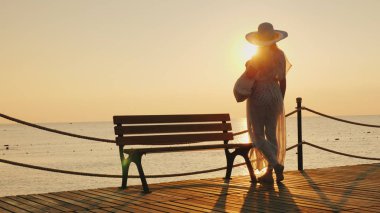 A pretty, slender girl in a developing pareo and a wide-brimmed stylish hat is standing on the pier, admiring the sunrise and enjoying the peace and tranquility. clipart