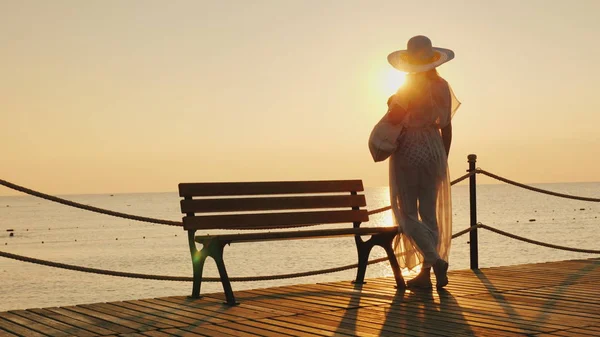 Ein hübsches, schlankes Mädchen in einem sich entwickelnden Pareo und einem breitkrempigen, stylischen Hut steht auf dem Pier, bewundert den Sonnenaufgang und genießt die Ruhe und Stille.. — Stockfoto