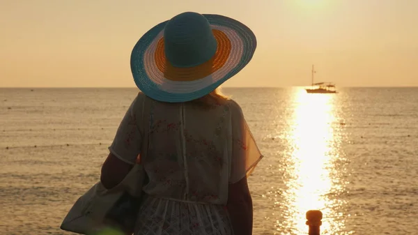 Uma jovem mulher com roupas de praia e um chapéu de abas largas está desfrutando do nascer do sol na praia. Vista traseira — Fotografia de Stock
