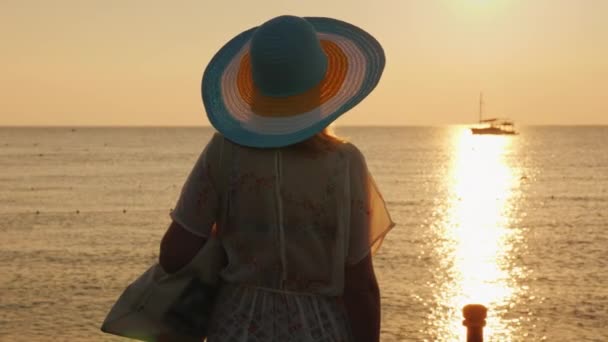 Plaj kıyafetleri ve geniş ekledi şapkası genç bir kadında gündoğumu sahil üzerinde keyif alıyor. Arkadan Görünüş — Stok video