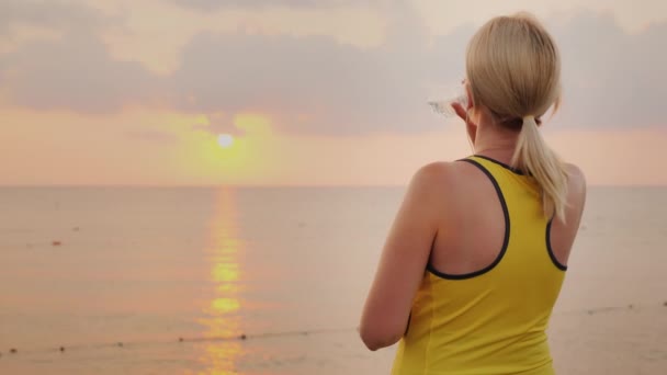 La mujer de fitness está bebiendo agua de una botella. Se encuentra en un lugar pintoresco junto al mar al amanecer. Concepto Agua Limpia y Salud — Vídeo de stock