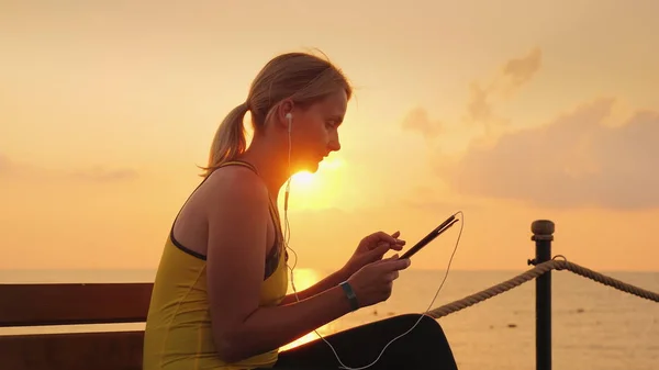 Mulher fitness goza de um tablet. Senta-se em um banco em um cais contra o pano de fundo do sol nascente — Fotografia de Stock